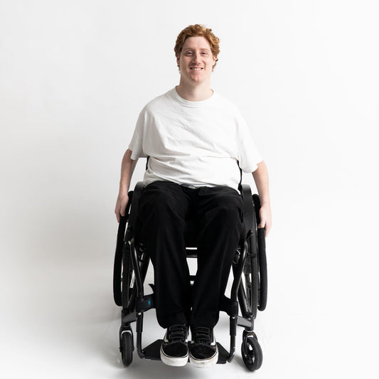 Model seated in a wheelchair wearing adaptive straight-leg pants in black with accessible pockets and elastic waistband.”