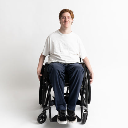 Model seated in a wheelchair wearing adaptive straight-leg pants in navy with accessible pockets and elastic waistband.