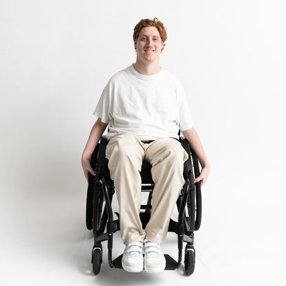 Model seated in a wheelchair wearing adaptive straight-leg pants in beige with accessible pockets and elastic waistband.

